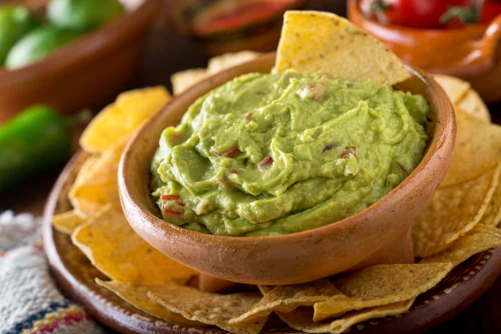 Guacamole dip with avocado, lime, and tomato surrounded by nacho chips.