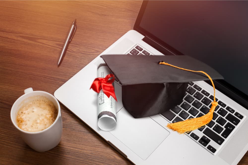 A cup of coffee along with a laptop topped with a graduation cap and diploma.